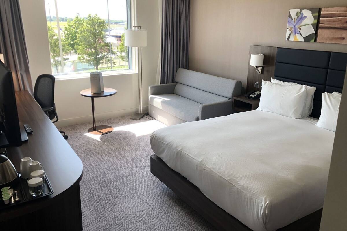 A hotel room with a bed, a desk, a chair, a sofa, a lamp, and a view of the outdoors.