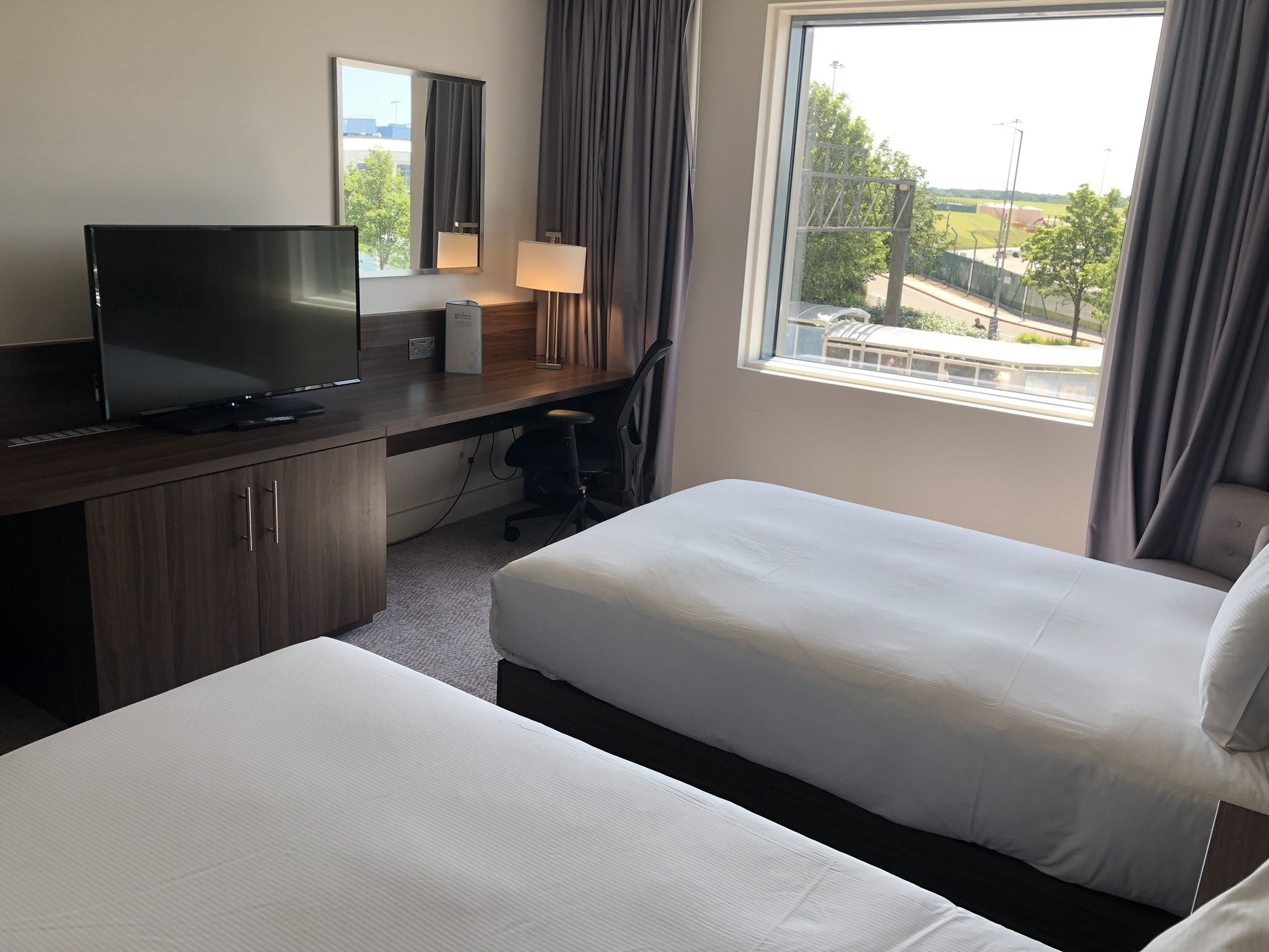 Habitación de hotel con escritorio, televisor de pantalla plana, dos camas individuales, ventana con vistas al exterior y lámpara.