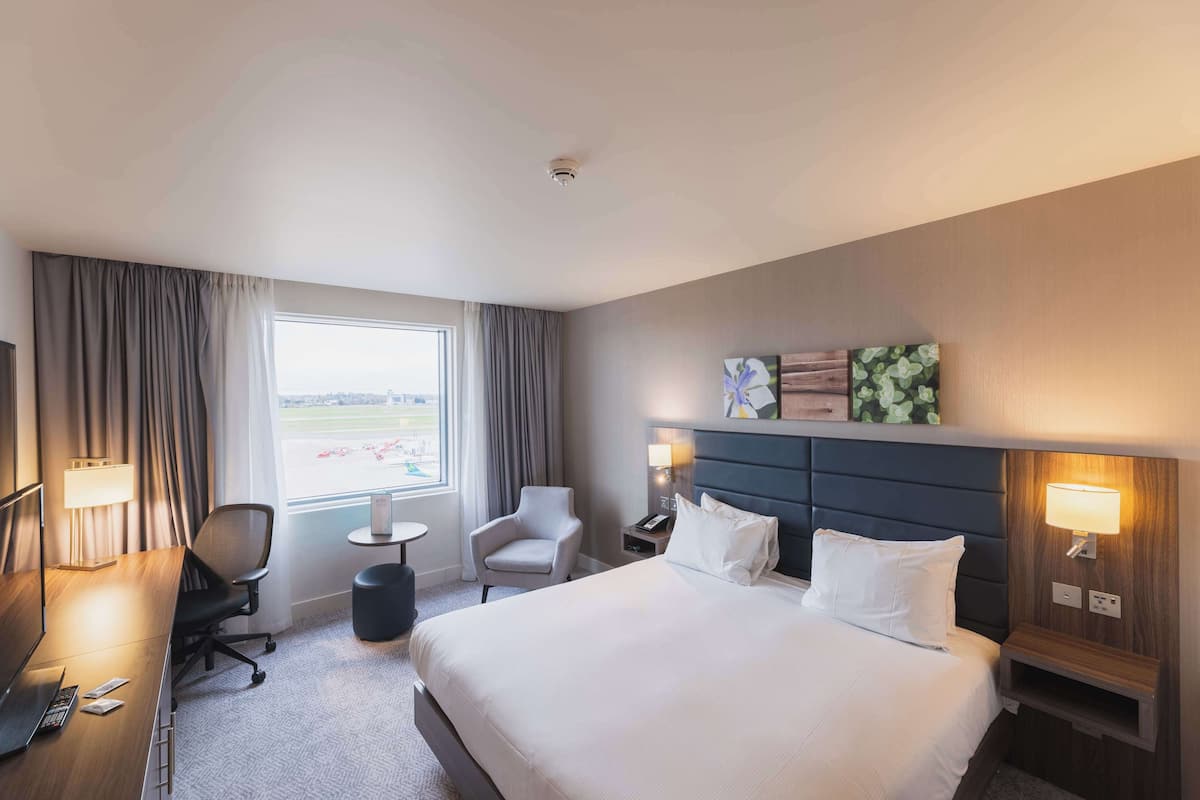 A hotel room with a large bed, a desk with a computer, a chair, a small table, and a window with curtains.