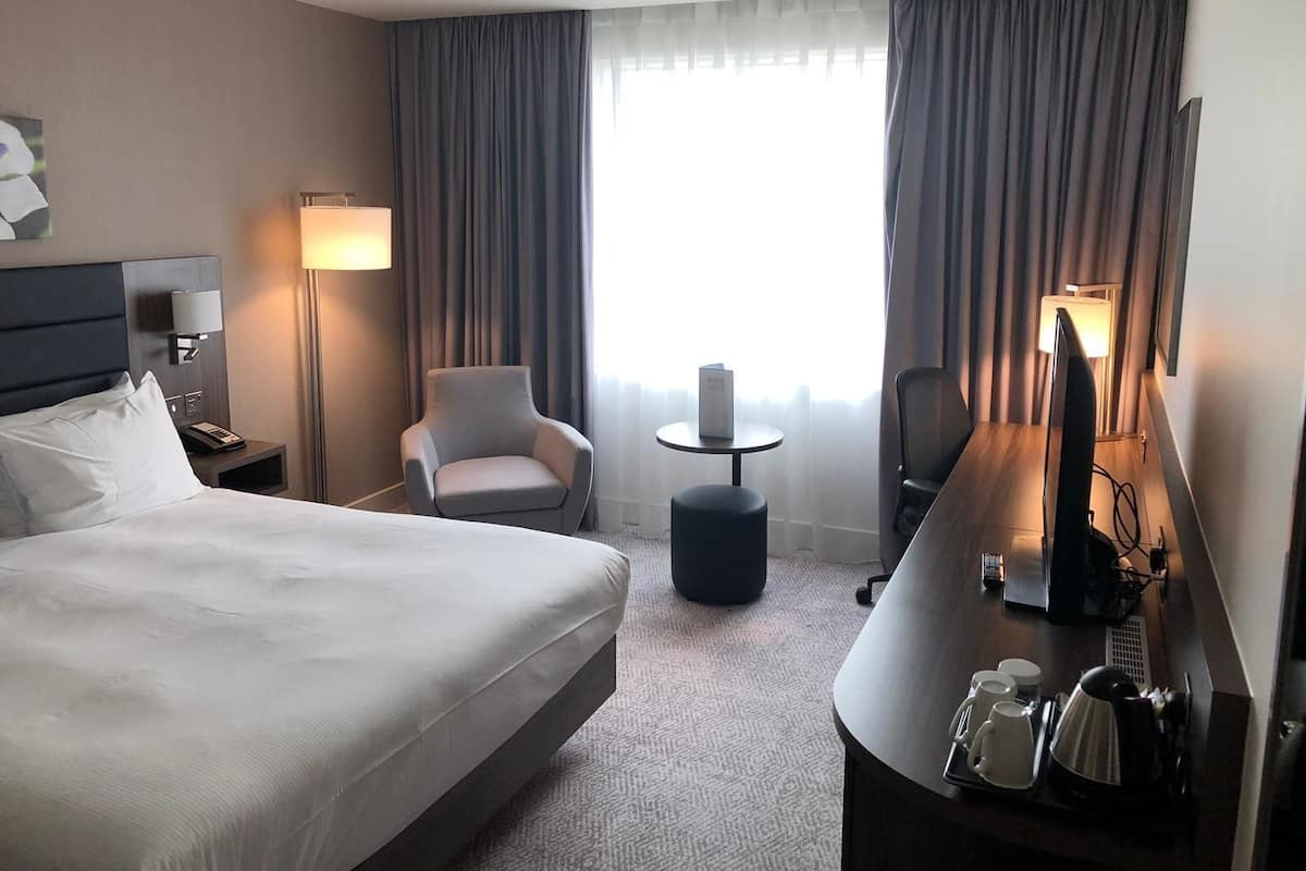 A hotel room with a bed, a desk with a coffee maker, a chair, a lamp, and a TV.