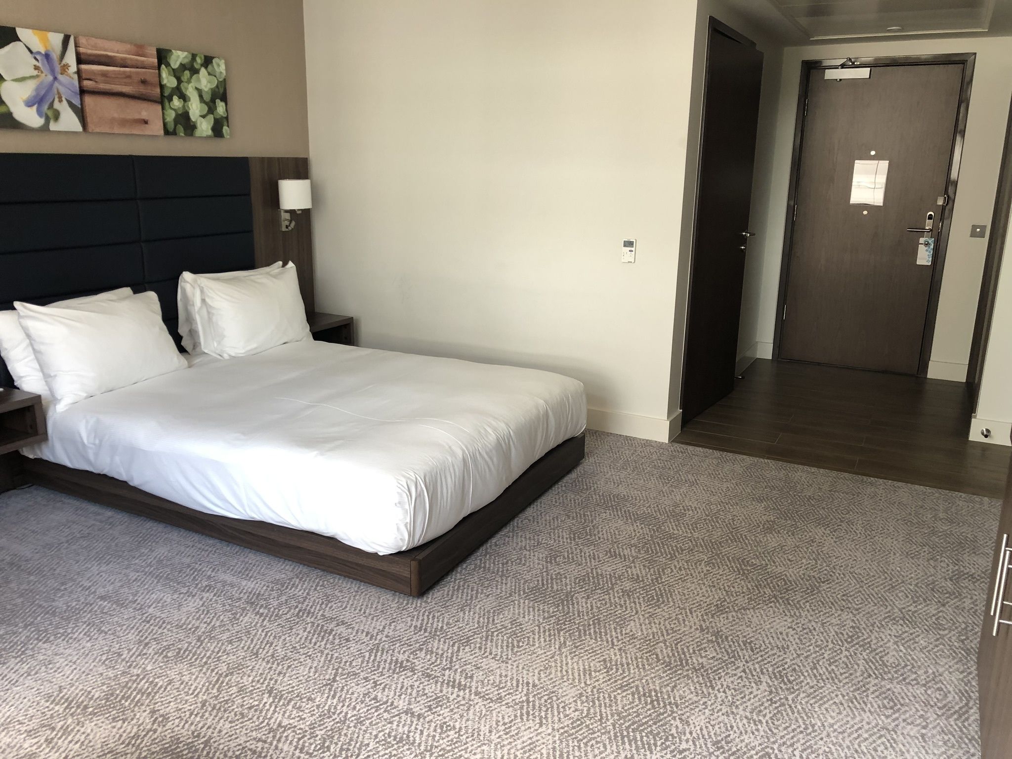 Una cama bien hecha en una habitación de hotel, con alfombra estampada, cabecero oscuro y una obra de arte en la pared.