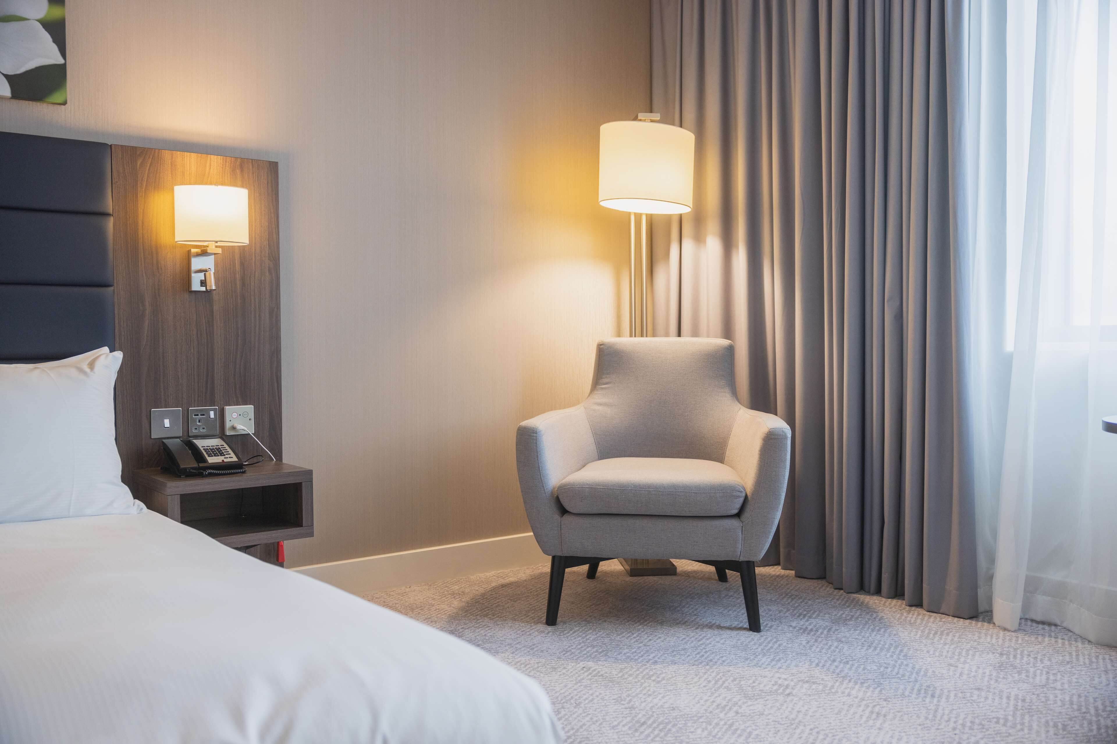 A hotel room with a bed, a nightstand with a phone, a lamp, and a chair.