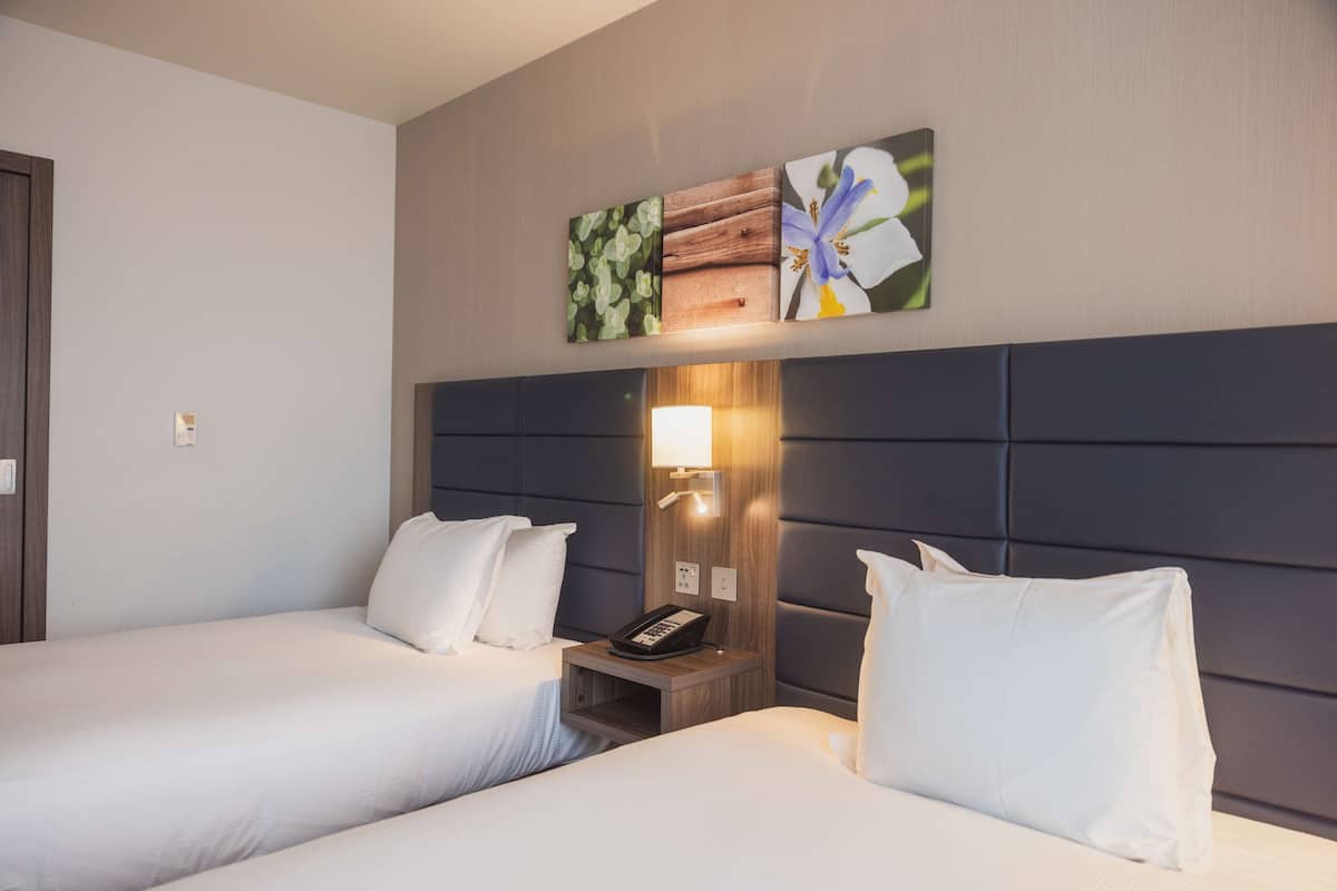 A hotel room with two beds, a nightstand with a lamp, and wall art above the beds.