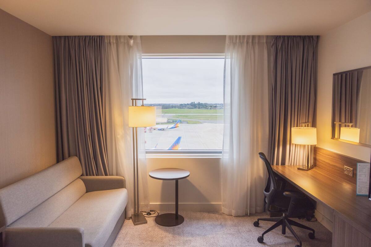 A hotel room with a sofa, a small round table, a desk with a chair, a lamp, and a window with a view of an airport.