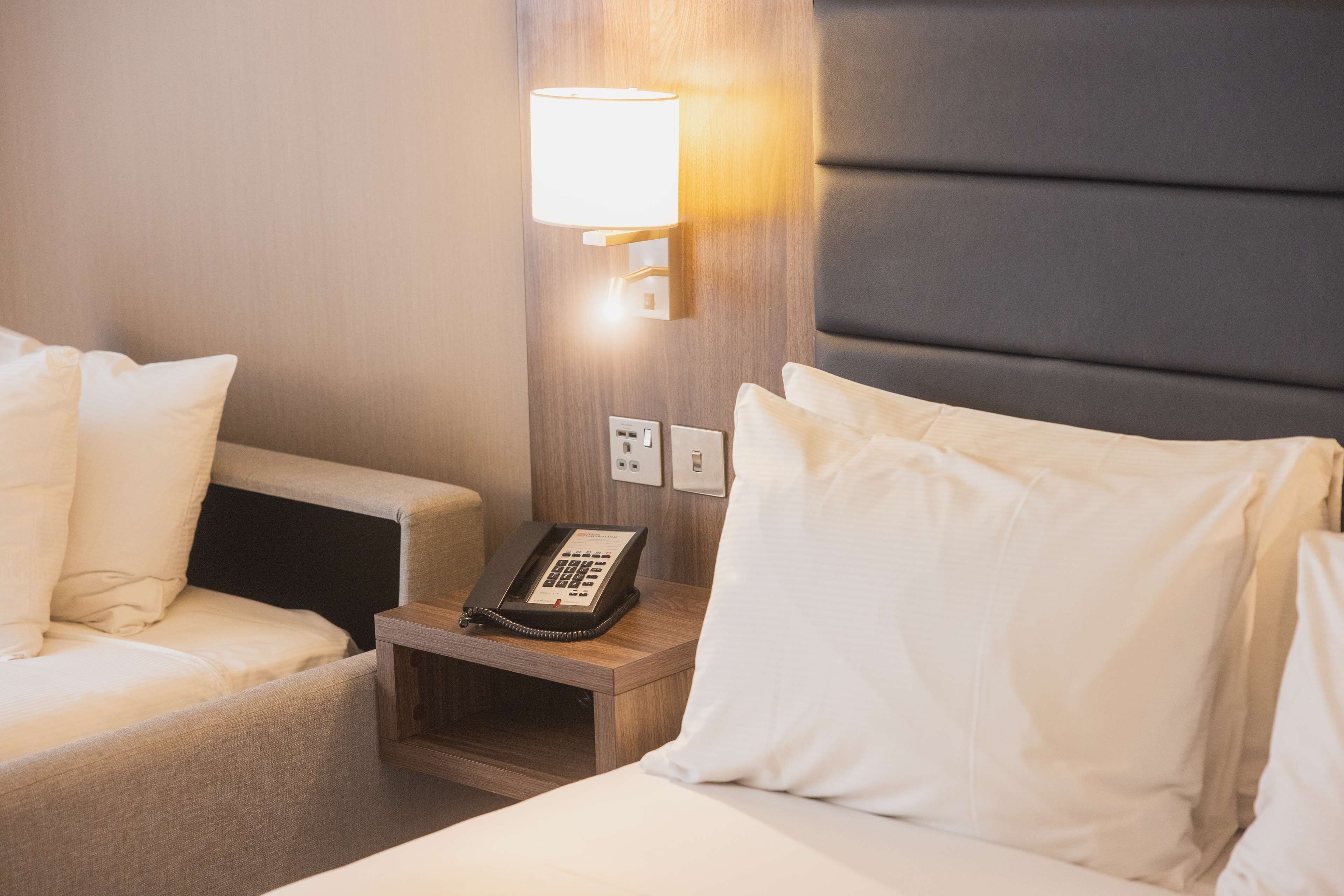 A hotel room with a bed, a bedside table with a phone, a lamp, and a wall socket.