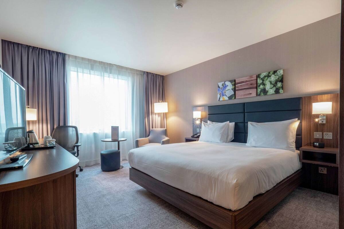 A hotel room with a large bed, a desk with a computer and phone, a chair, a small table, and a lamp.