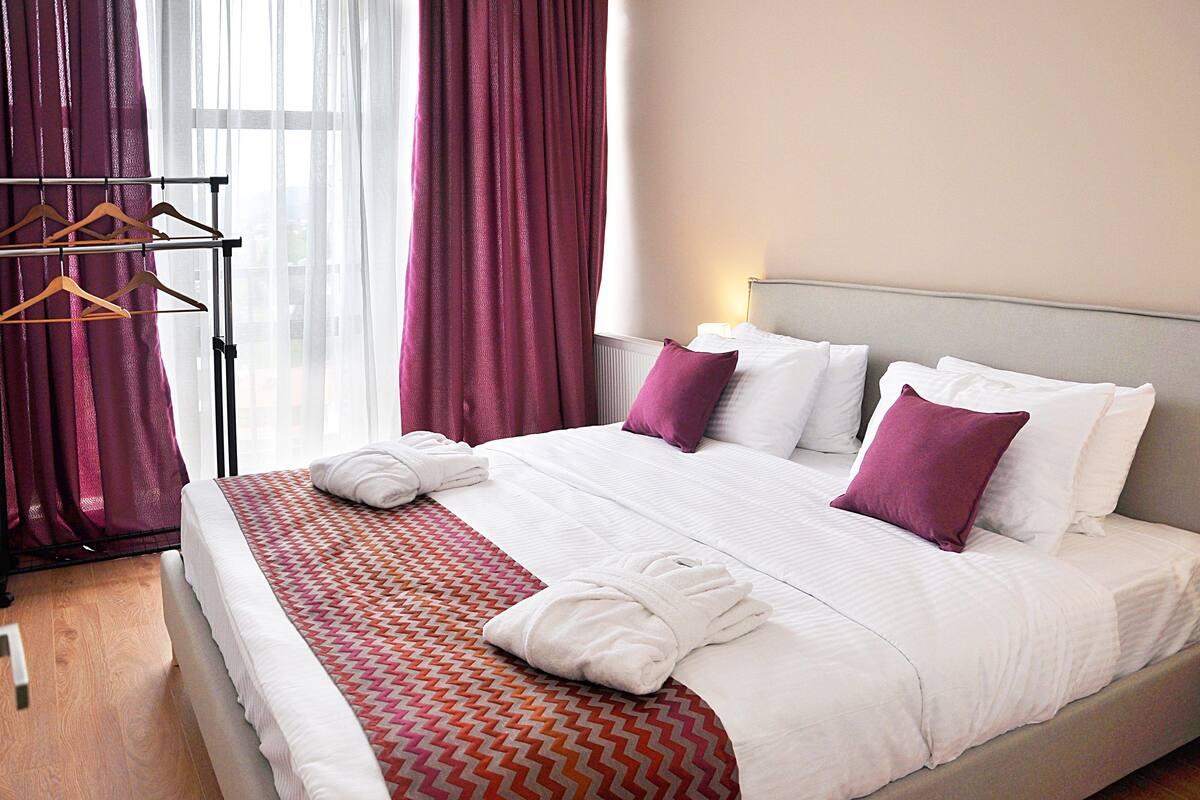 Una cama bien hecha con sábanas blancas y una colcha de cuadros rojo y naranja, una mesita de noche con lámpara y una ventana con cortinas blancas traslúcidas y un cortinaje morado.