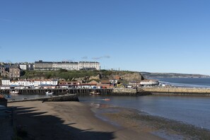 A coastal town with a prominent building on a hill, a pier with boats, and a sandy beach.