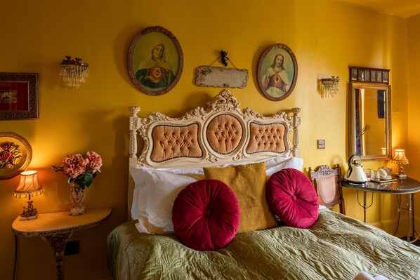 A bed with a tufted headboard, two red pillows, a green bedspread, and a bedside table with a lamp and flowers.