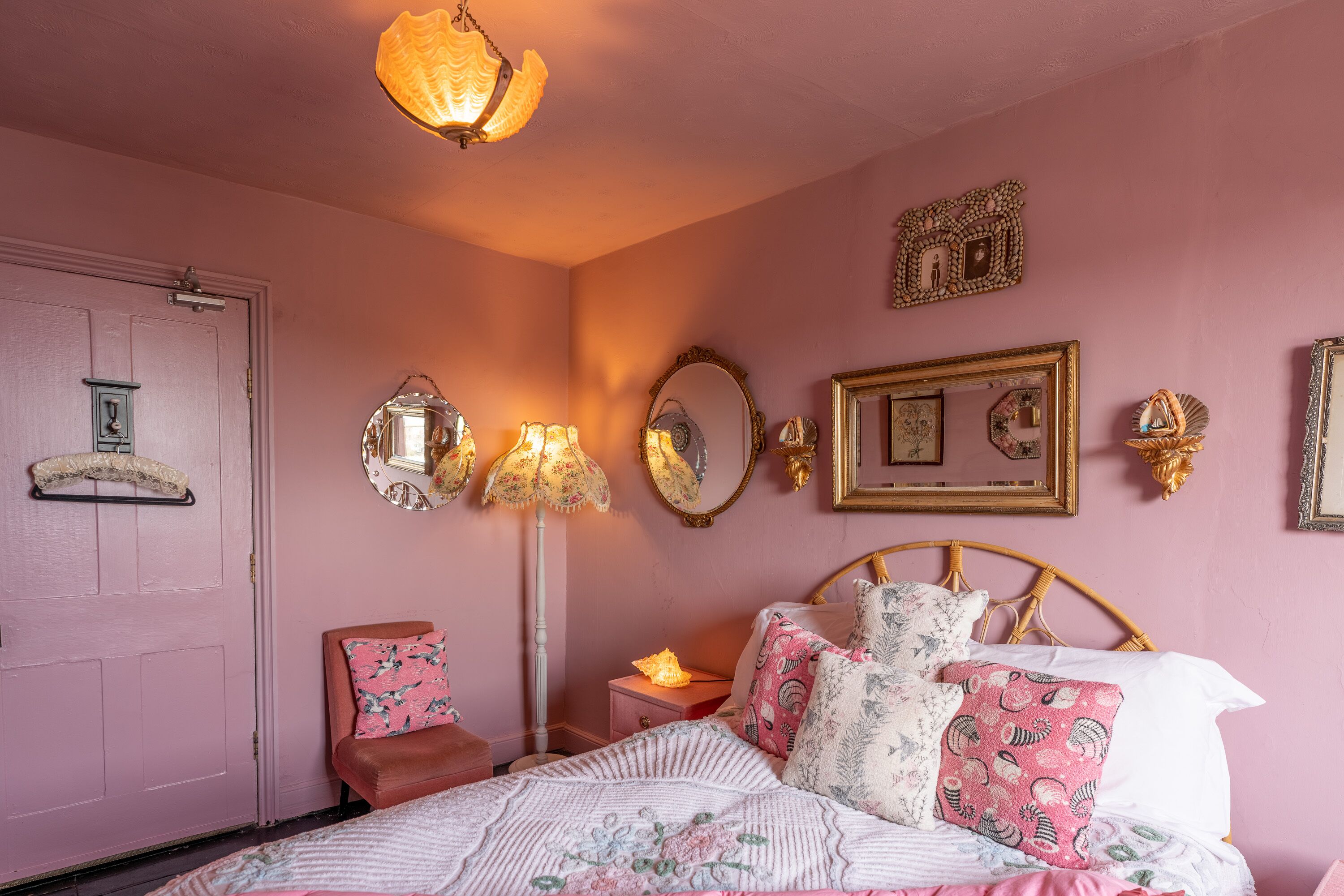 Een slaapkamer in roze tinten, met een bed, kussens, een nachtkastje, een lamp en wanddecoraties.