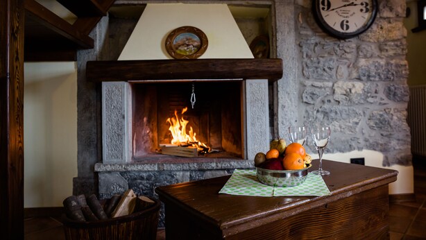 Una chimenea de piedra con un reloj encima, una mesa de madera con un frutero y vasos, y un alero de la chimenea con un cuadro.