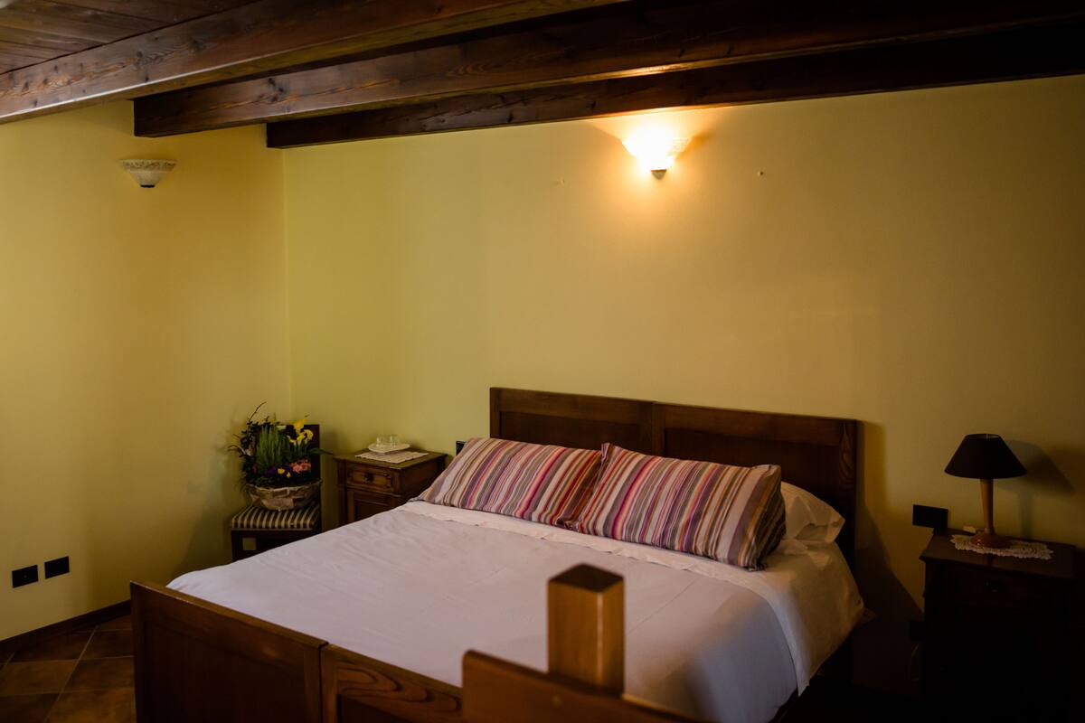 A neatly made bed with striped pillows, a bedside lamp, a nightstand with a lamp, and a small table with a plant and a telephone.