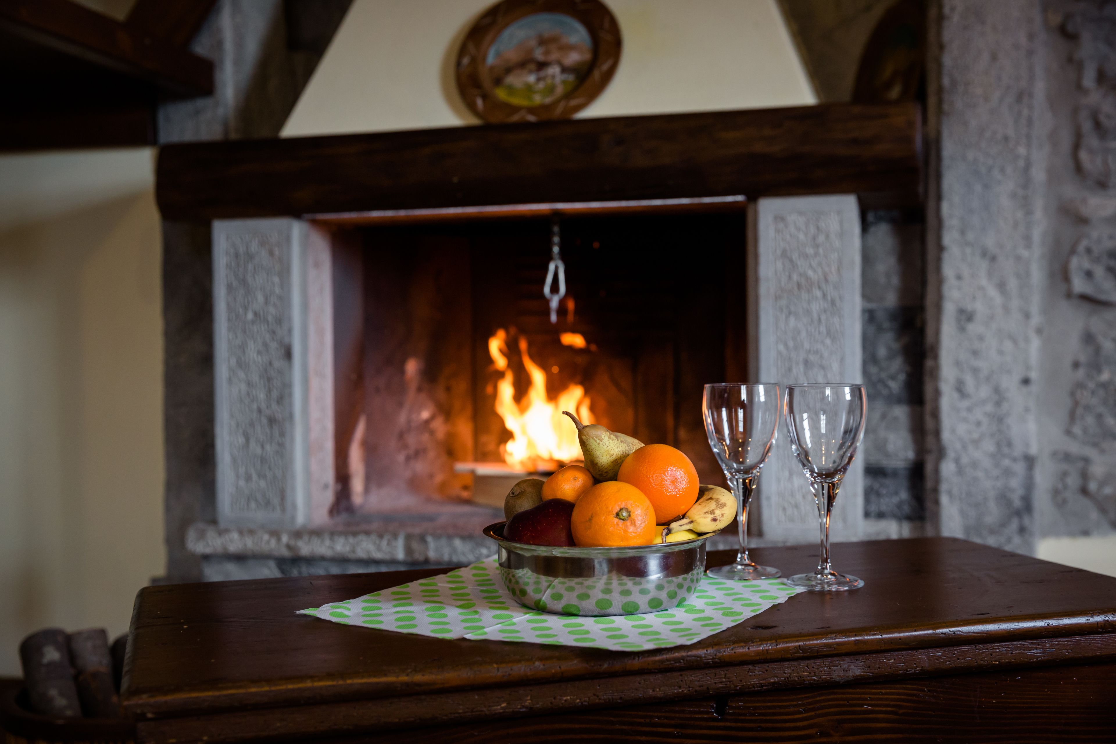 A wooden table with a fruit bowl and two wine glasses. A fireplace with a burning fire is in the background.