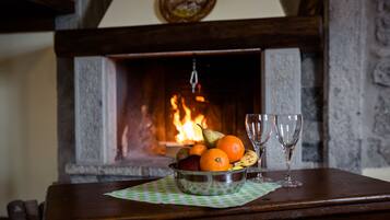 A wooden table with a fruit bowl and two wine glasses. A fireplace with a burning fire is in the background.