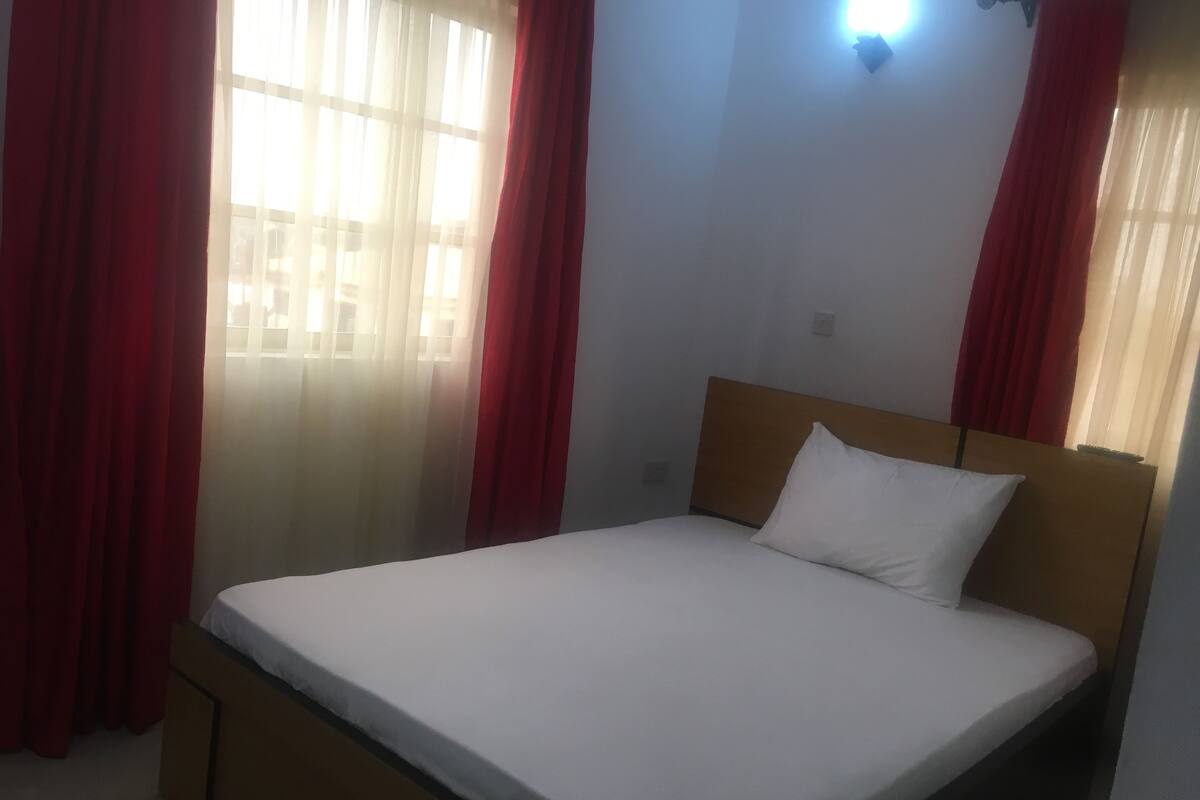 A single bed with a wooden headboard in a room with red curtains and a window with sheer curtains.