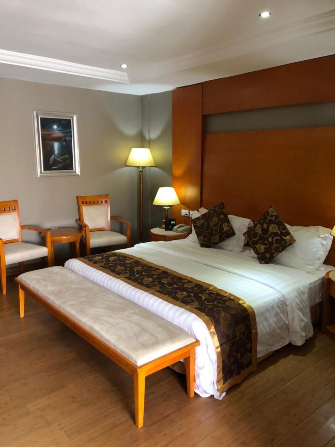 A hotel room with a bed, a bench, two chairs, a nightstand, a lamp, and a painting on the wall.