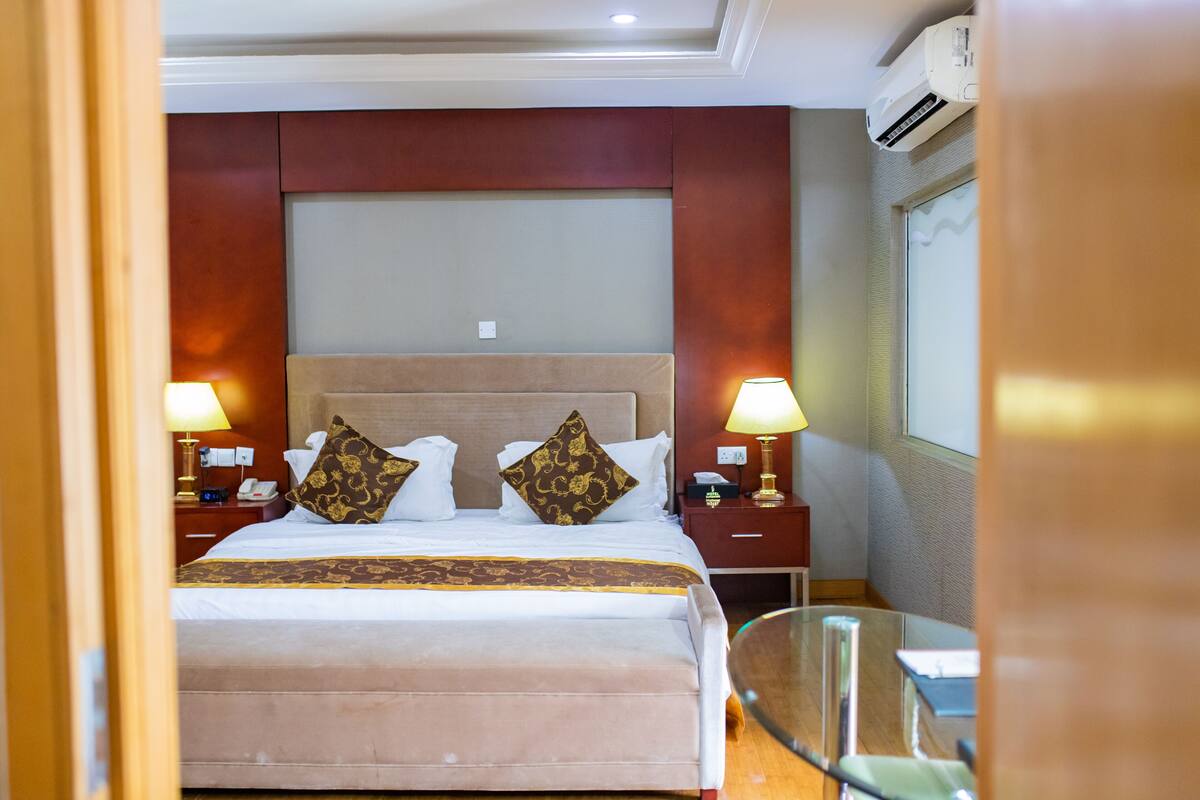 A hotel room with a large bed, two bedside tables with lamps, a glass table, and an air conditioning unit.