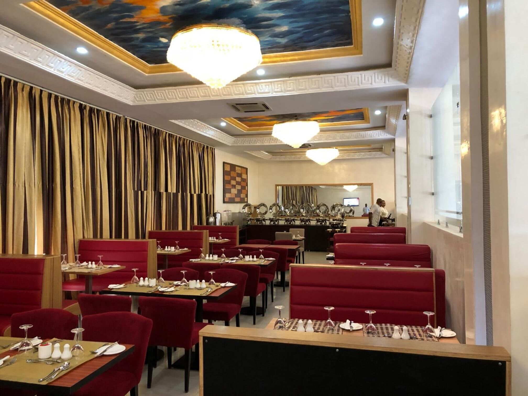 Un restaurante con mesas y puestos tapizados en rojo, una araña y un mural en el techo.