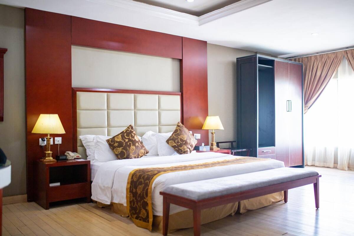 A hotel room with a large bed, two bedside tables with lamps, a bench, a wardrobe, and a large window with curtains.