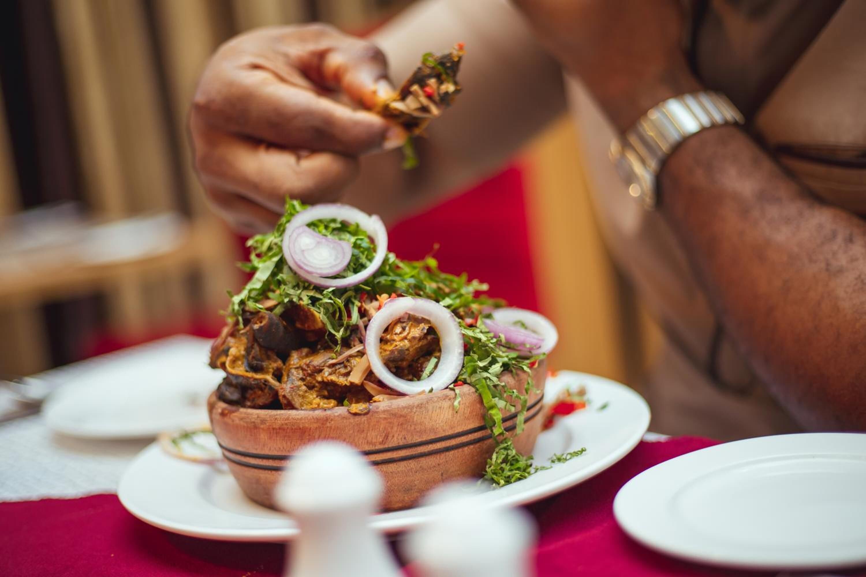 A person serving food from a wooden bowl on a table with a red tablecloth.