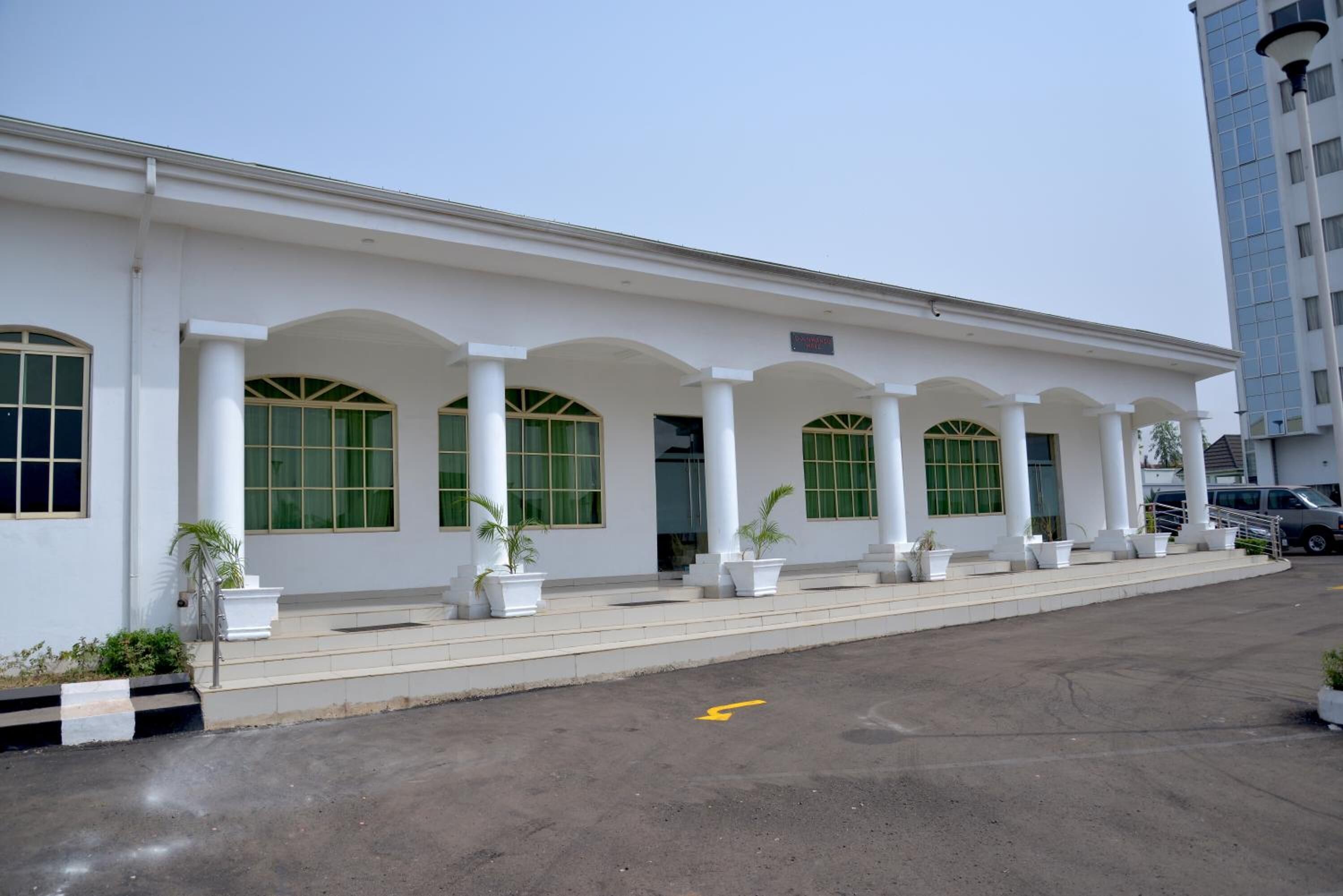 A white building with a row of arched windows and columns, a paved entrance area with potted plants, and a clear sky.