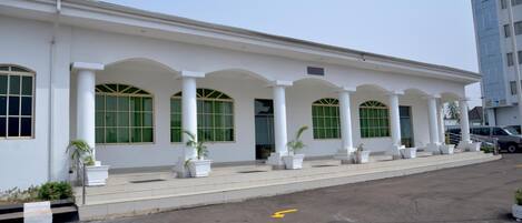 A white building with a row of arched windows and columns, a paved entrance area with potted plants, and a clear sky.
