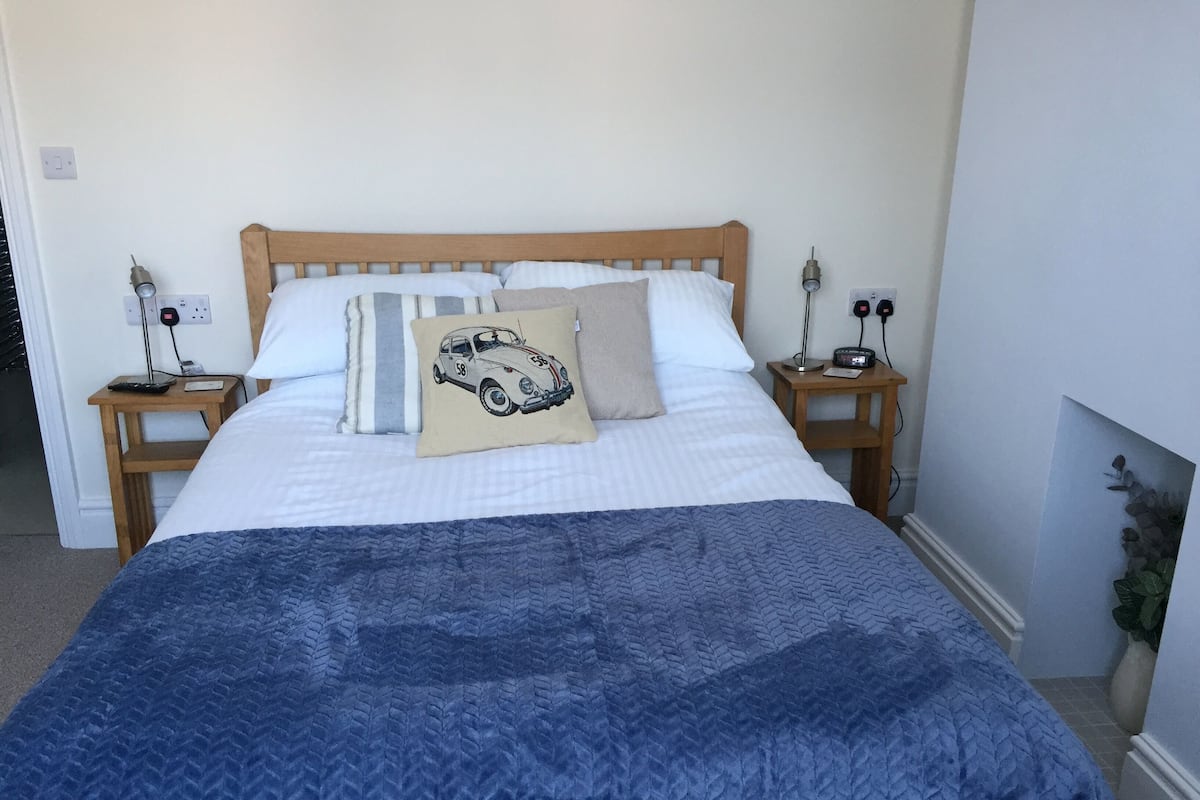 Cama bem arrumada com manta azul, duas mesas de cabeceira com abajures e luminária na parede.