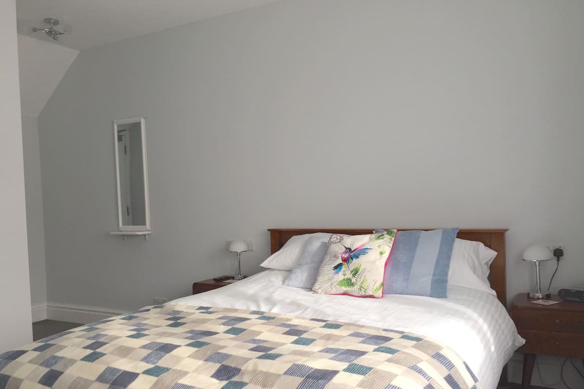 Quarto com colcha xadrez, cabeceira de madeira, duas mesas de cabeceira com abajures e um espelho na parede.