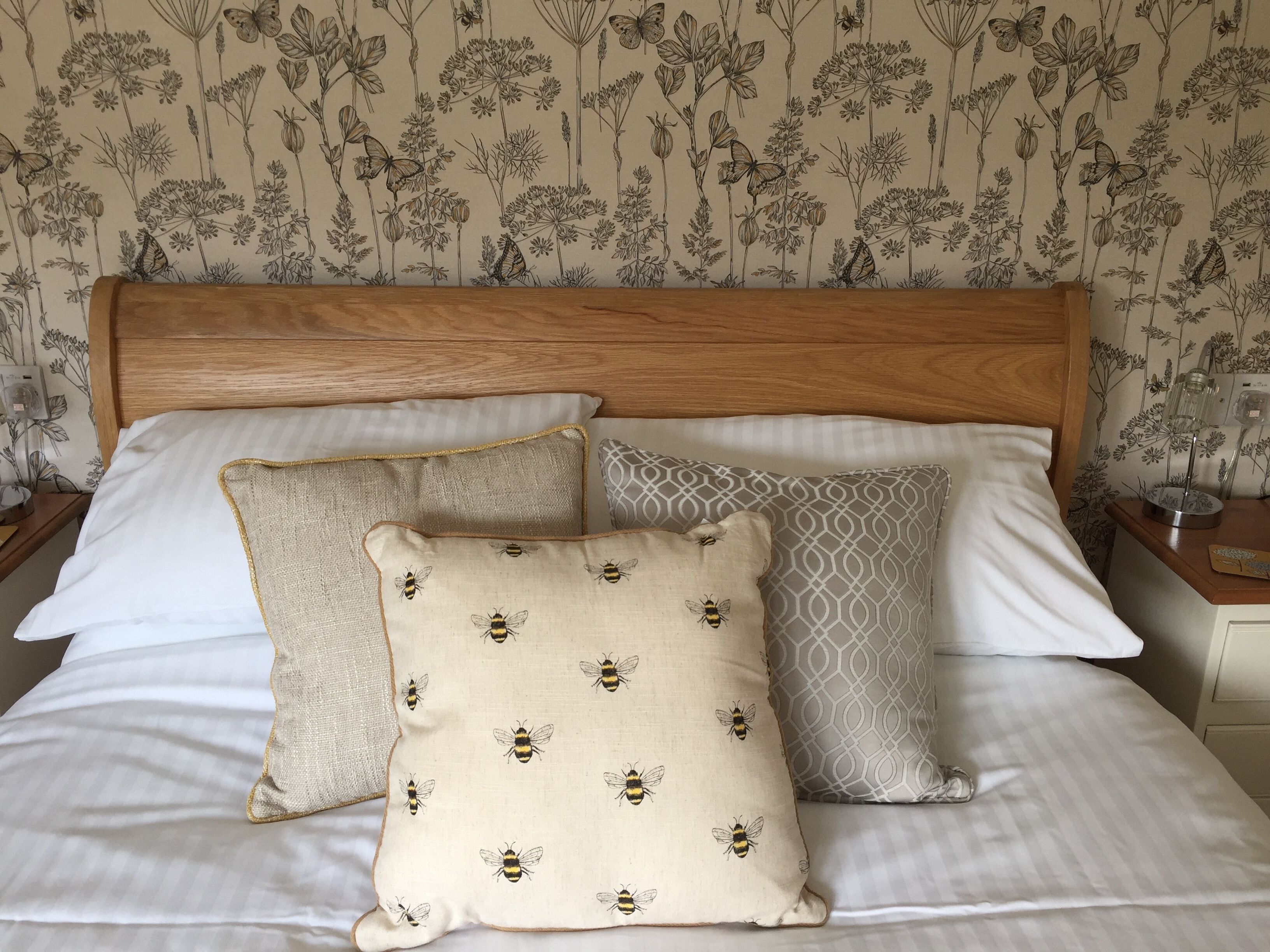 A neatly made bed with three pillows, one with a bee pattern, and a floral wallpaper background.