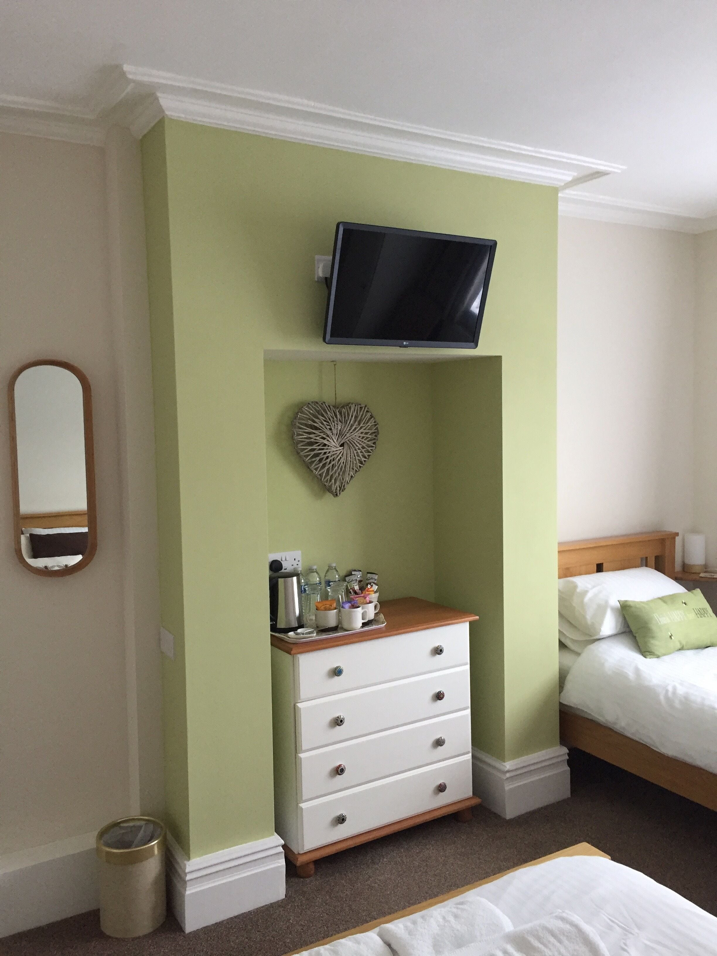 A bedroom with a bed, a dresser, a television mounted on the wall, a heart-shaped decoration, and a trash can.