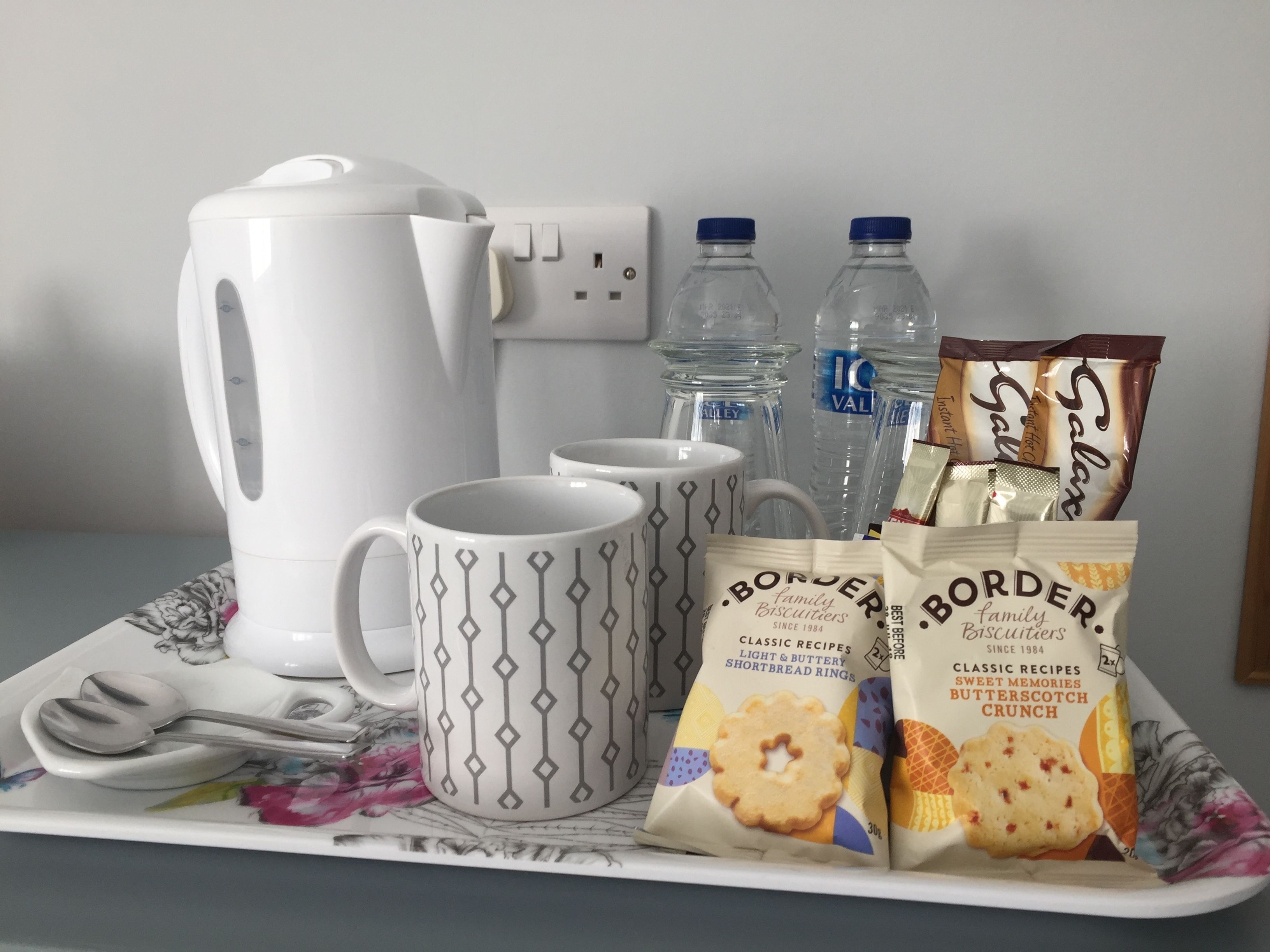 Een dienblad met een waterkoker, twee mokken, twee waterflessen en zakjes koekjes.