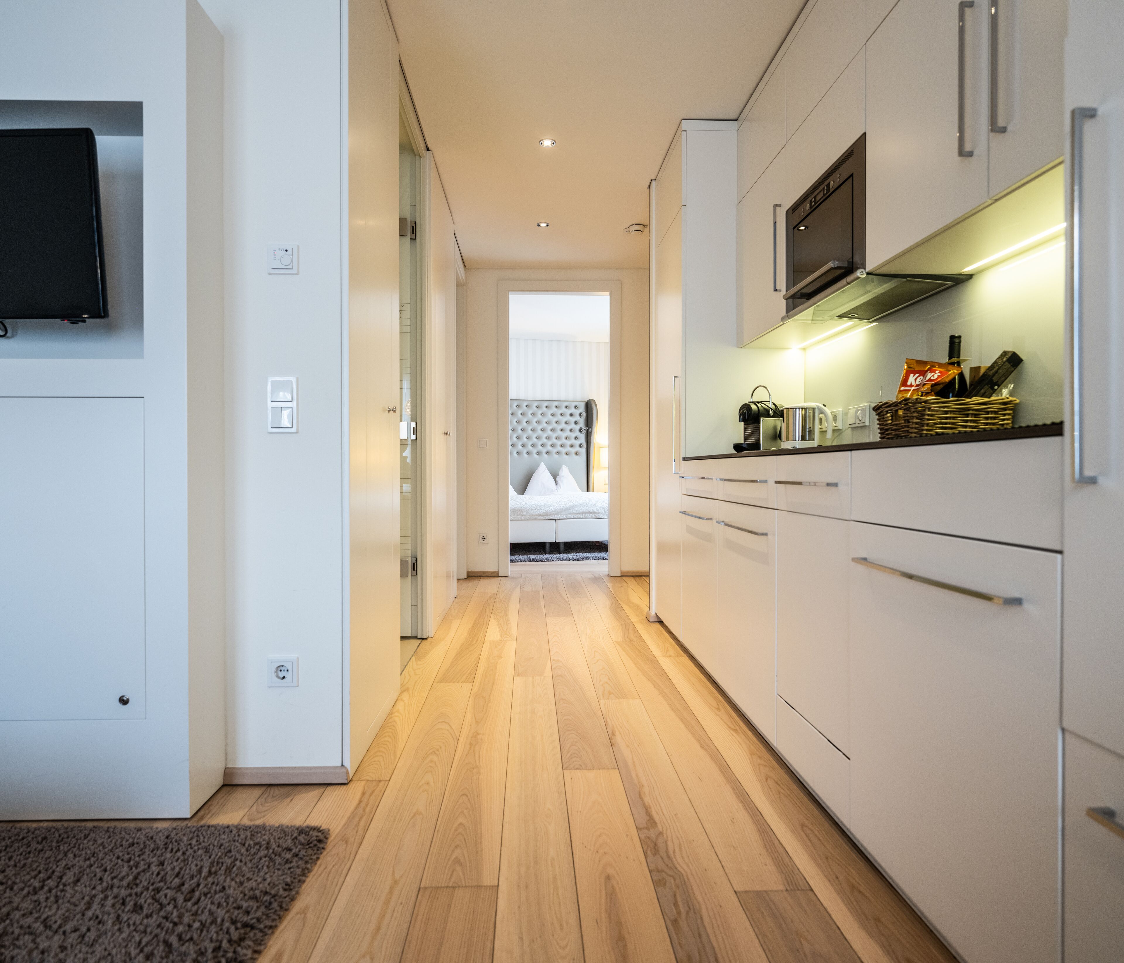 Una cocina moderna con armarios blancos, piso de madera y televisor de pantalla plana montado en la pared.