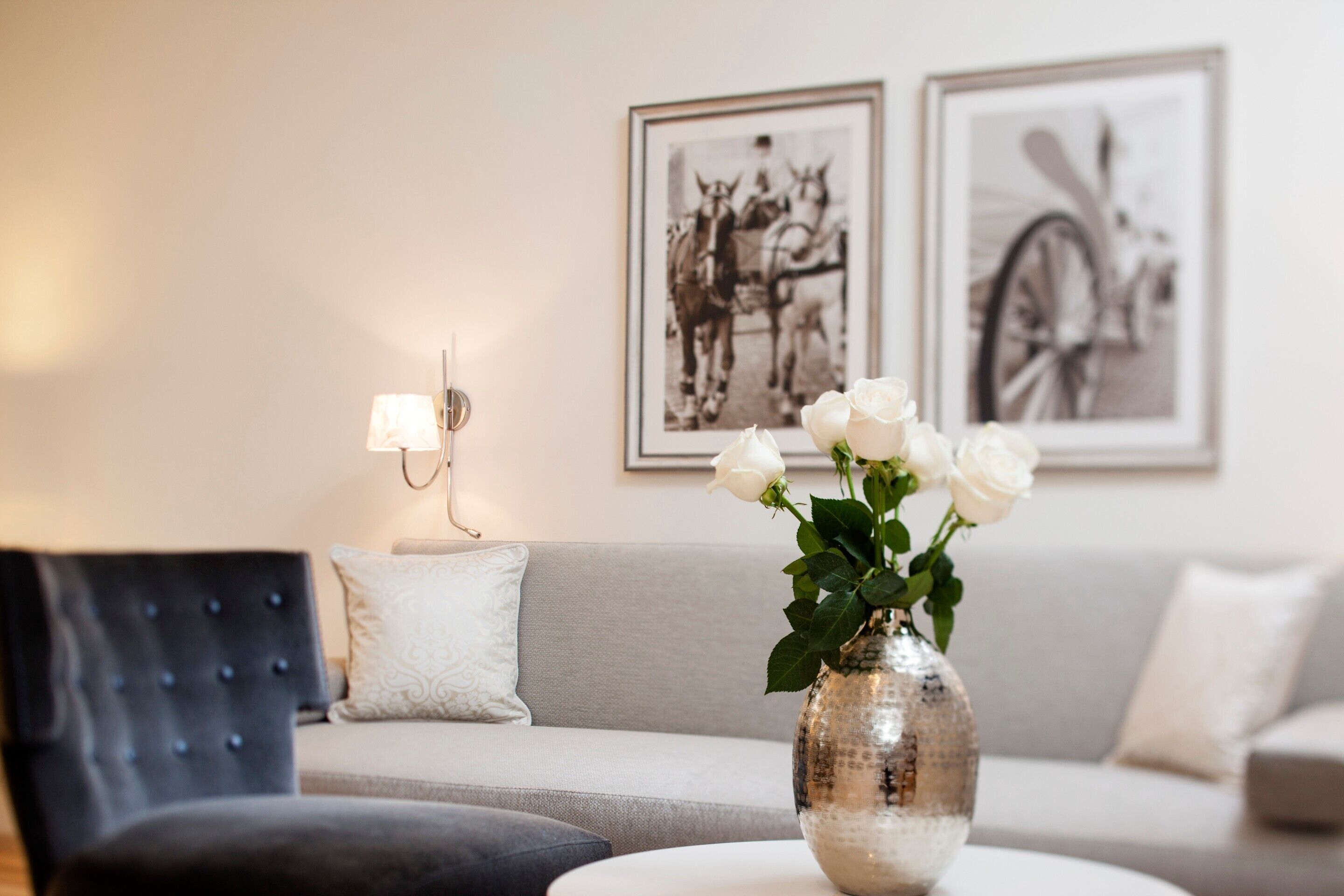 Una sala de estar con un sofá, una silla, un florero con rosas blancas y dos cuadros en la pared.