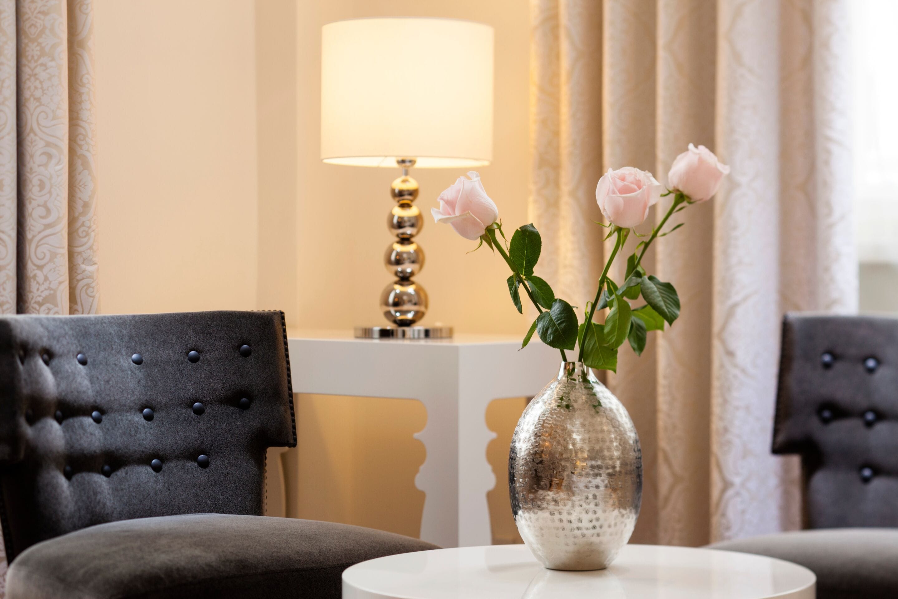 Una habitación con un sillón capitonado, una mesa redonda con un jarrón de rosas y una lámpara.