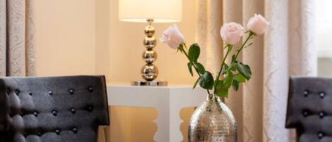 A room with a tufted armchair, a round table with a vase of roses, and a lamp.