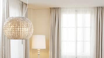 A modern hotel room with a dining area, two upholstered chairs, a round table, a chandelier, and a large window with curtains.
