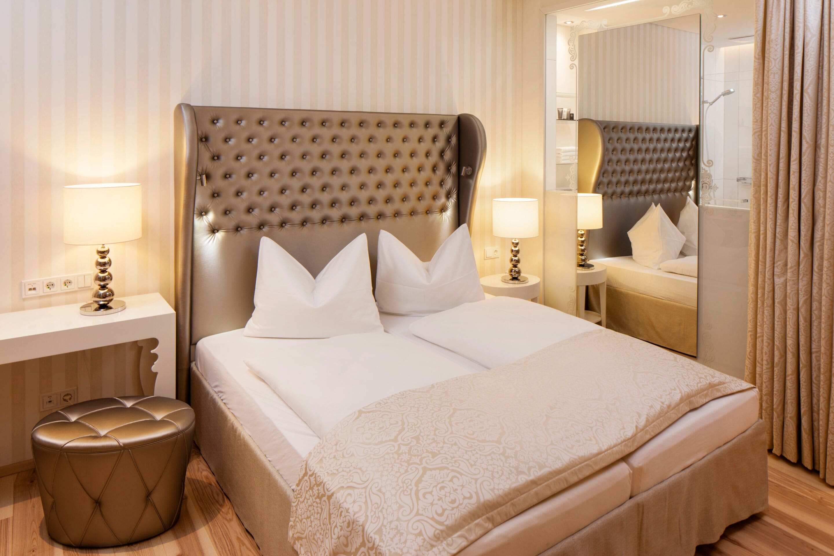 A neatly made bed with a tufted headboard, two bedside tables with lamps, a round ottoman, and a mirror reflecting the room.