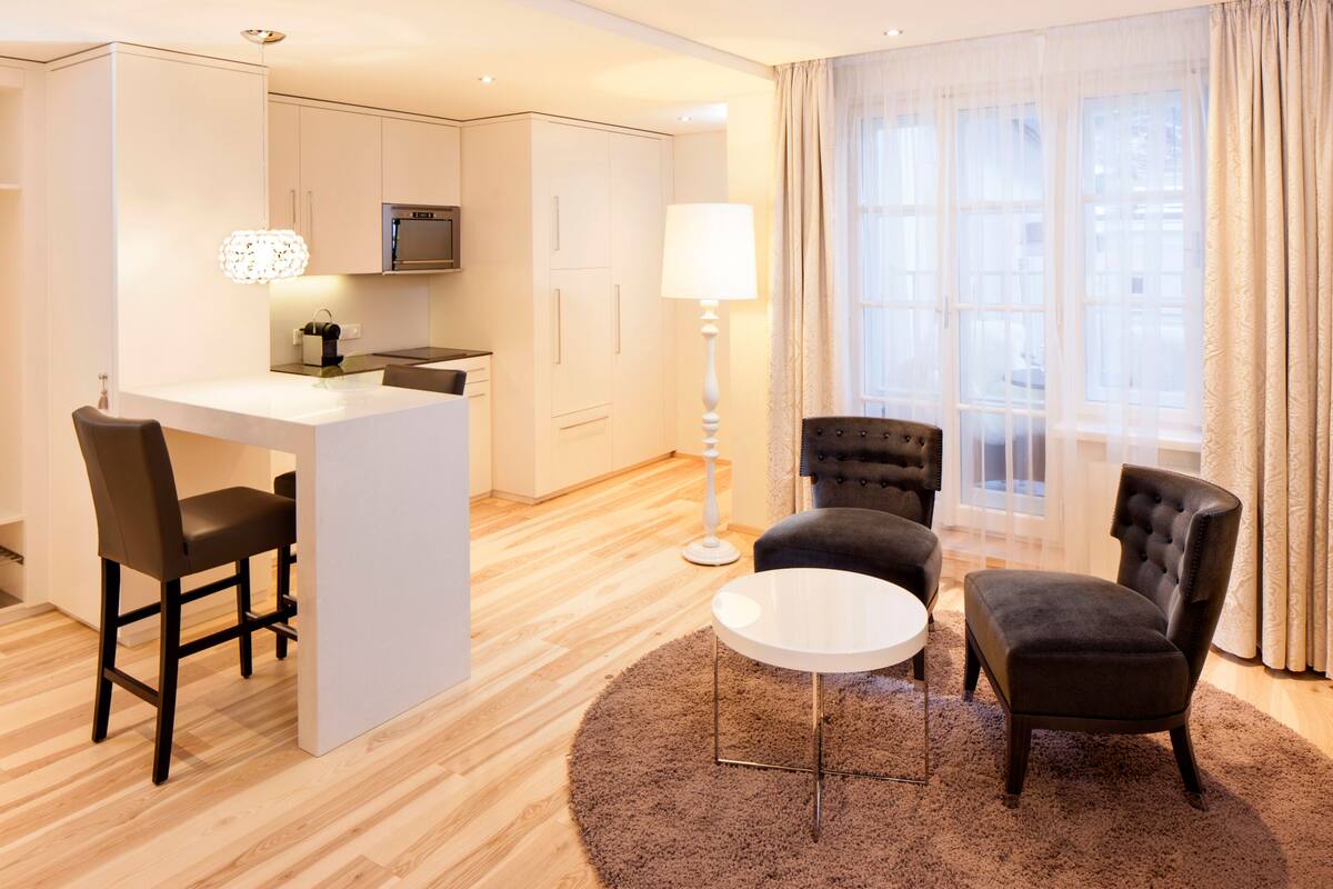 Une chambre d’hôtel moderne dotée d’une kitchenette, d’un coin repas et d’un espace salon avec deux fauteuils et une petite table ronde.