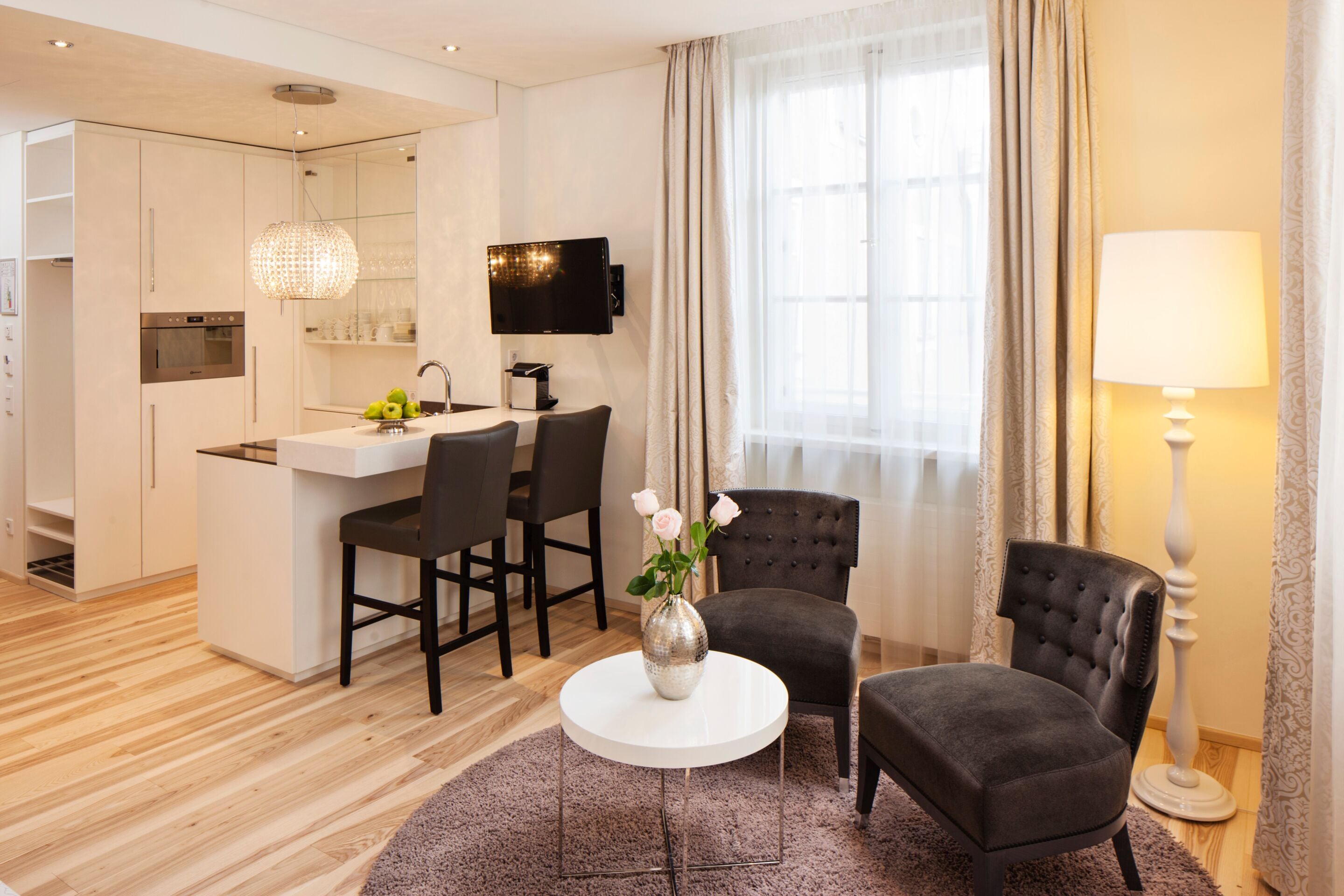 A modern hotel room with a kitchen area, a dining table with chairs, a sofa, and a lamp.