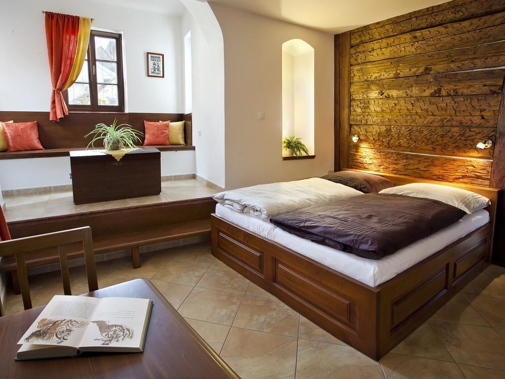 A bedroom with a wooden headboard, a bed with white and brown bedding, a wooden bench with cushions, a small table with a book, and a window with curtains.