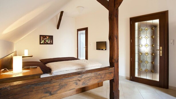 A bedroom with a sloped ceiling, a wooden bed frame, a bedside table with a lamp, and a door with a frosted glass panel. - Staroměstský penzion (Tabor)