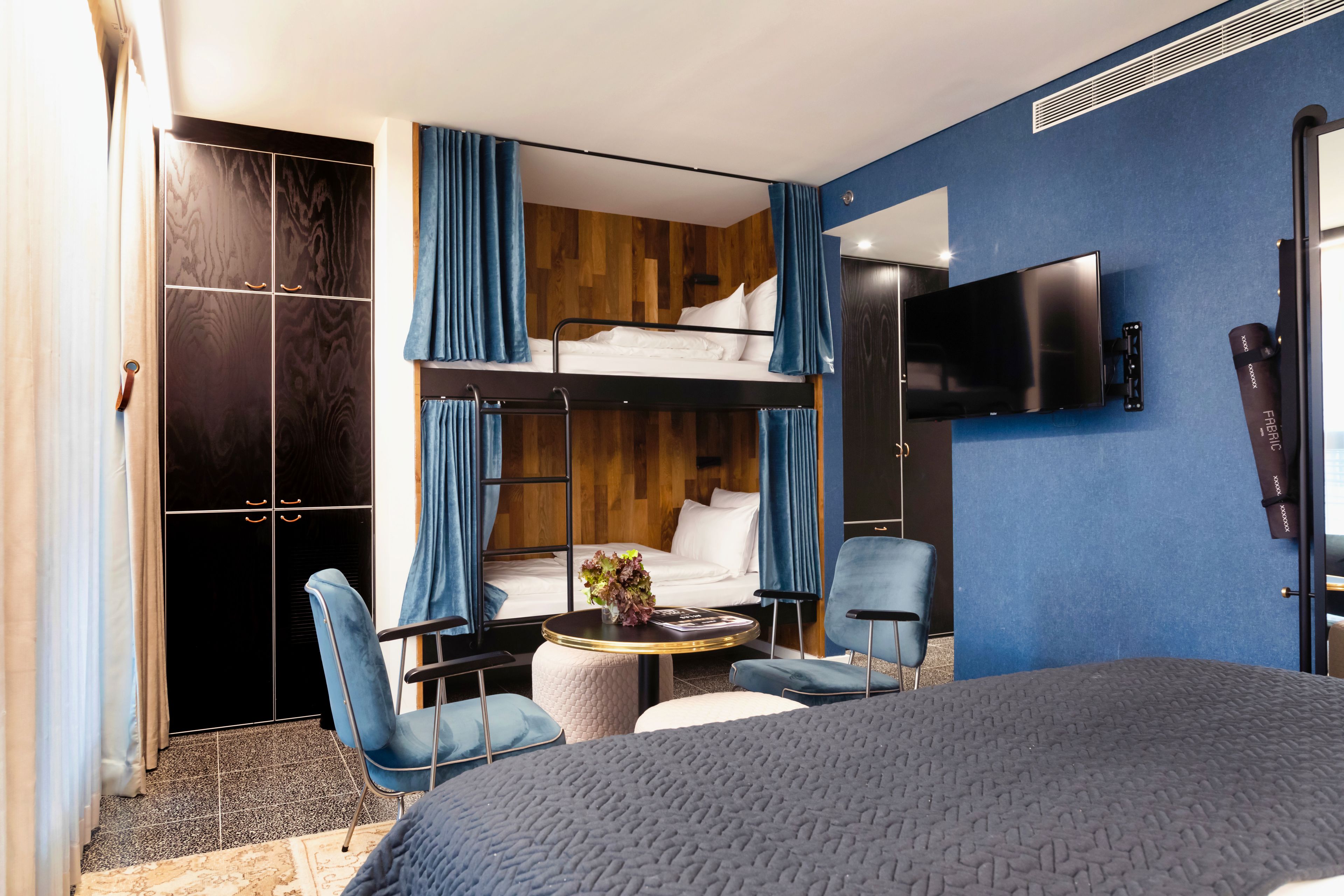 A hotel room with a bunk bed, a flat-screen TV, a round table with a vase, and a blue chair.