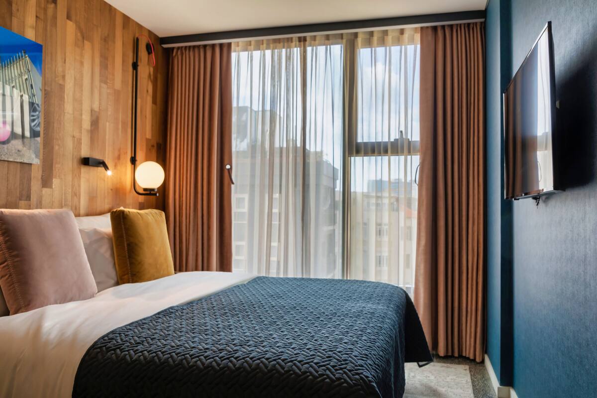 Una habitación de hotel con cama, televisión en la pared y ventana con cortinas.