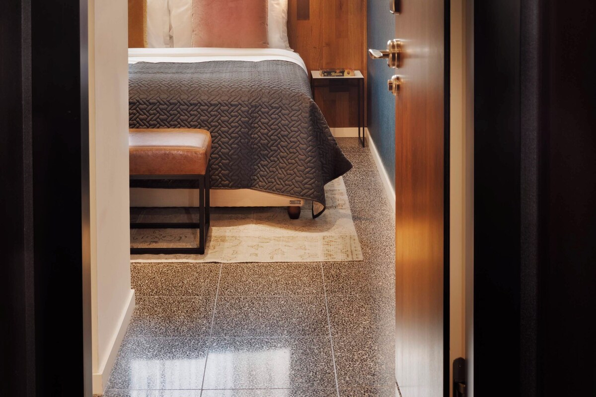 Een hotelkamer met een bed, een bankje, een tafeltje en een deur.