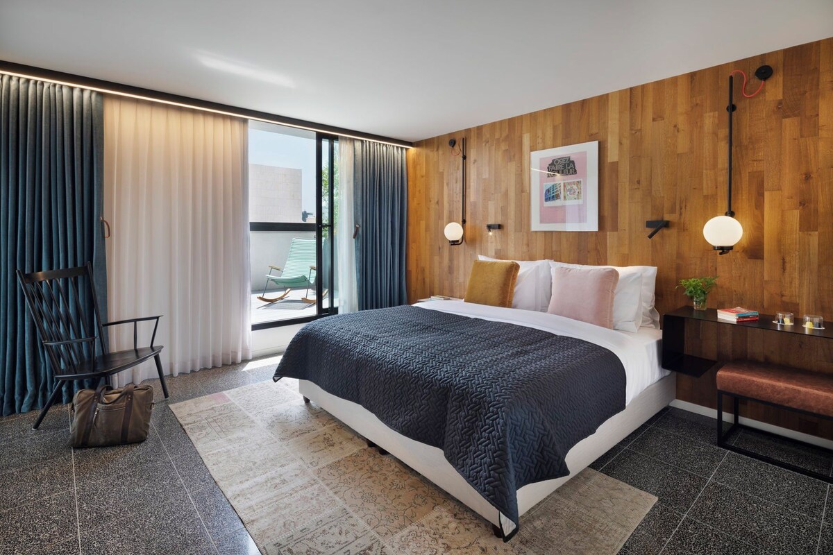 A modern bedroom with a large bed, a wooden accent wall, a desk with a chair, and a view of the outdoors.