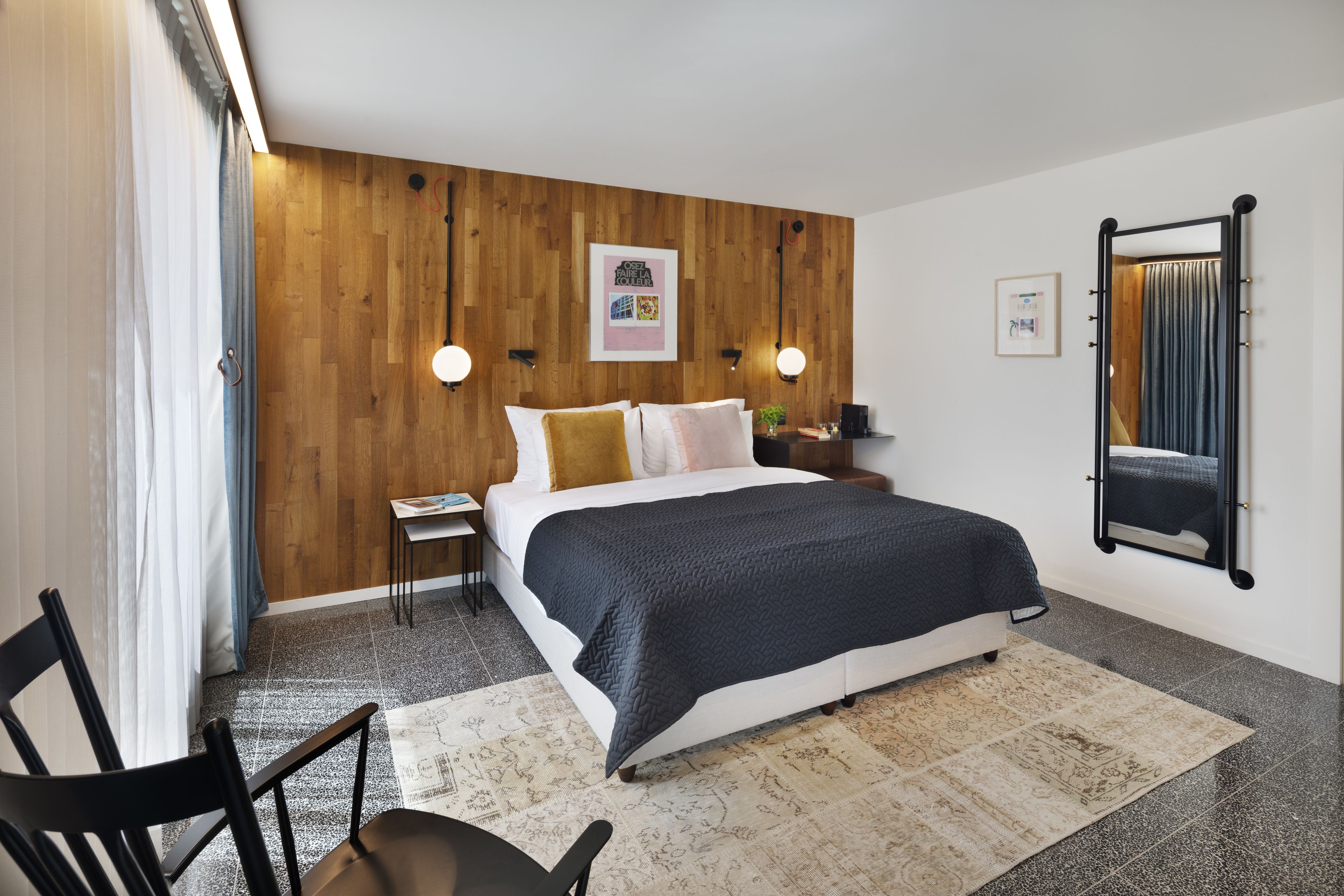 Un dormitorio con panel de madera, una cama con manta oscura, dos mesitas de noche con lámparas, un espejo y un cuadro enmarcado en la pared.