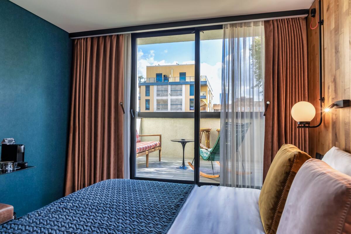 Una habitación de hotel con una cama grande, un balcón con mesa y sillas, y vistas a edificios.