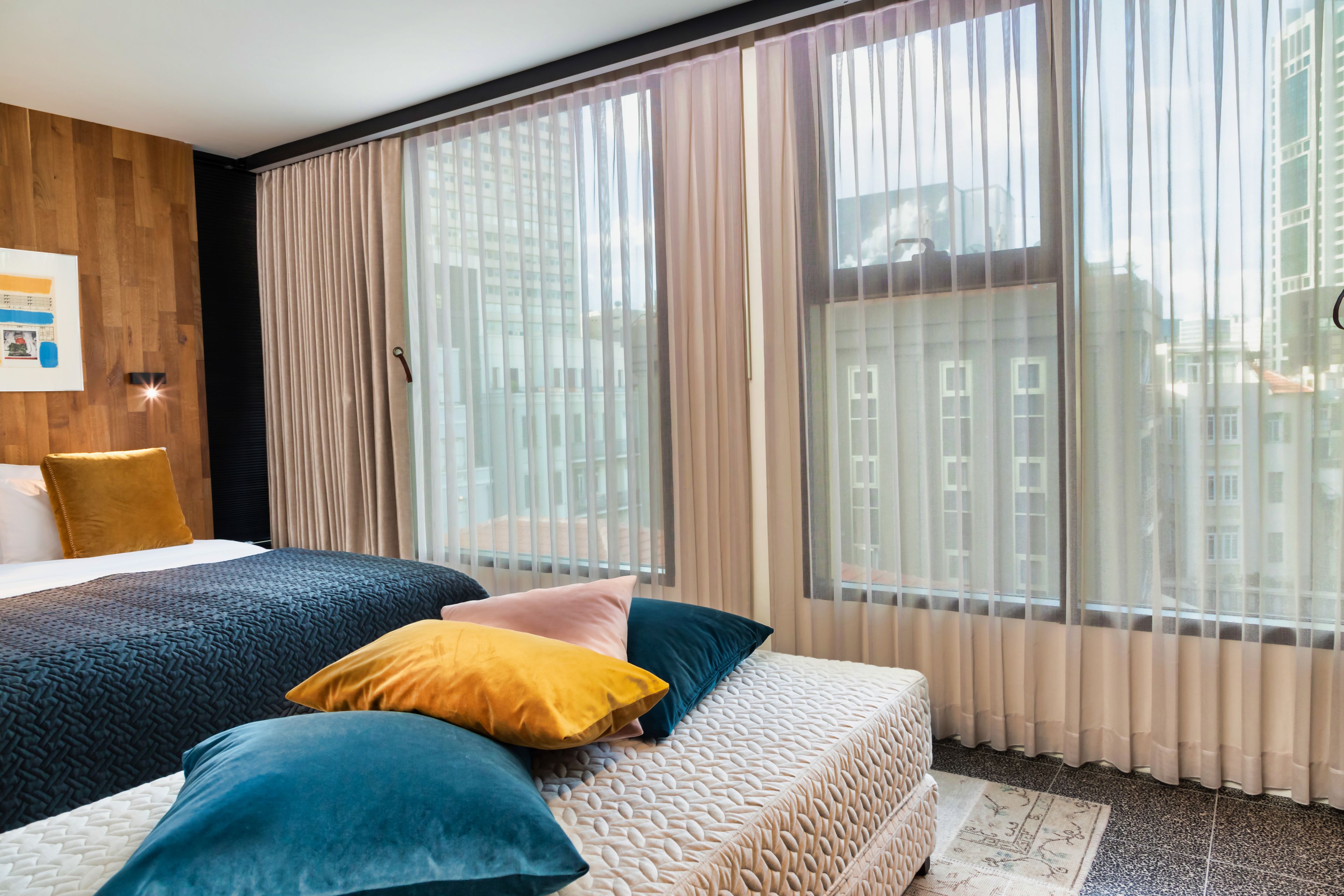Habitació d'hotel moderna amb un llit gran, una paret decorativa de fusta i vista a la ciutat a través de cortines translúcides.