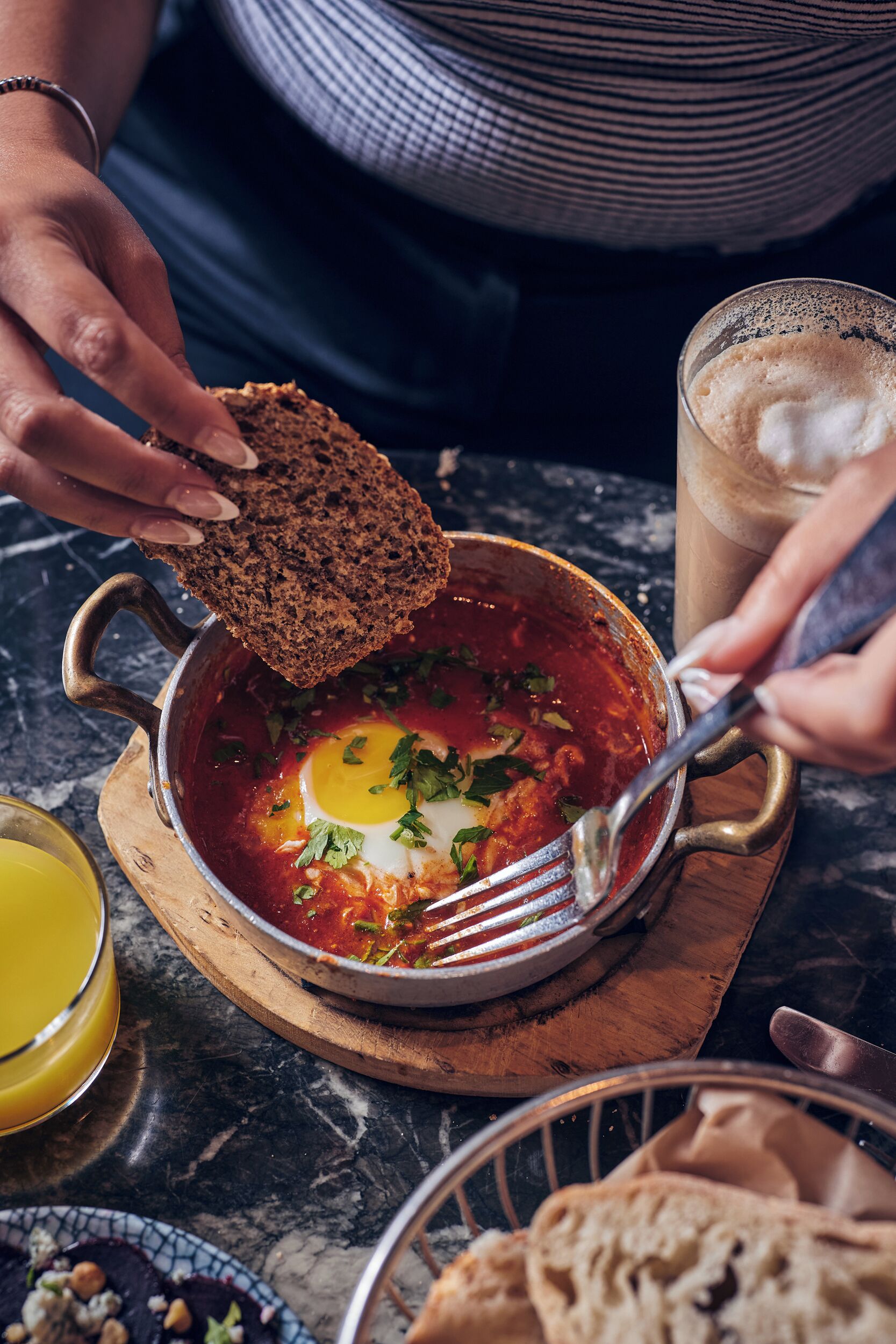 A person serving a dish with eggs and sauce, a glass of iced coffee, and a bowl of bread on a wooden board.