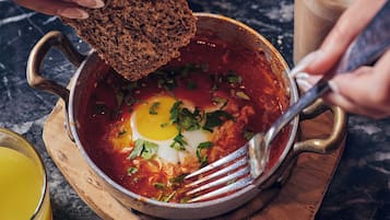A person serving a dish with eggs and sauce, a glass of iced coffee, and a bowl of bread on a wooden board.